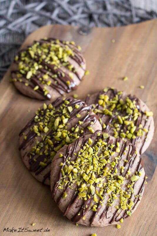Schokoladencookies mit Pistazien 1 Vier Schokoladen-Pistazien-Cookies auf einem Brettchen mit Schokostreifen und gehackten Pistazien verziert