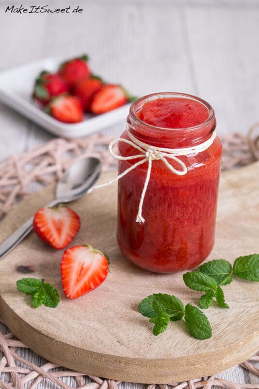 Fruchtige Erdbeer-Minze-Marmelade im Glas mit einer Schleife. Daneben liegen Erdbeeren, Minze und ein Löffel